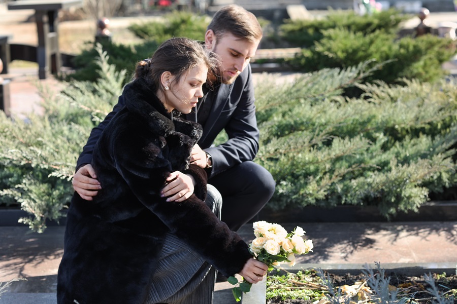 Frau hockt vor einem Grab und legt Blumen nieder. Ein Mann hockt neben ihr und umarmt sie seitlich.