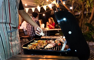 Grill mit Fleisch als Grillgut. Jemand in Grillschürze steht davor mit einer Grillzange.