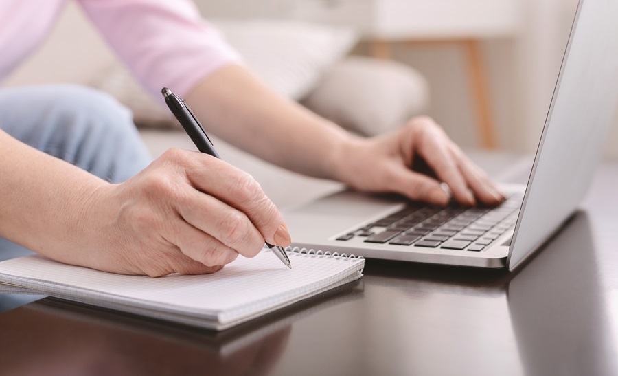 Laptop auf Tisch, daneben ein Block; die linke Hand einer Frau liegt auf der Tastatur des Laptops, die rechte Hand hält einen Stift, um auf dem Block zu schreiben